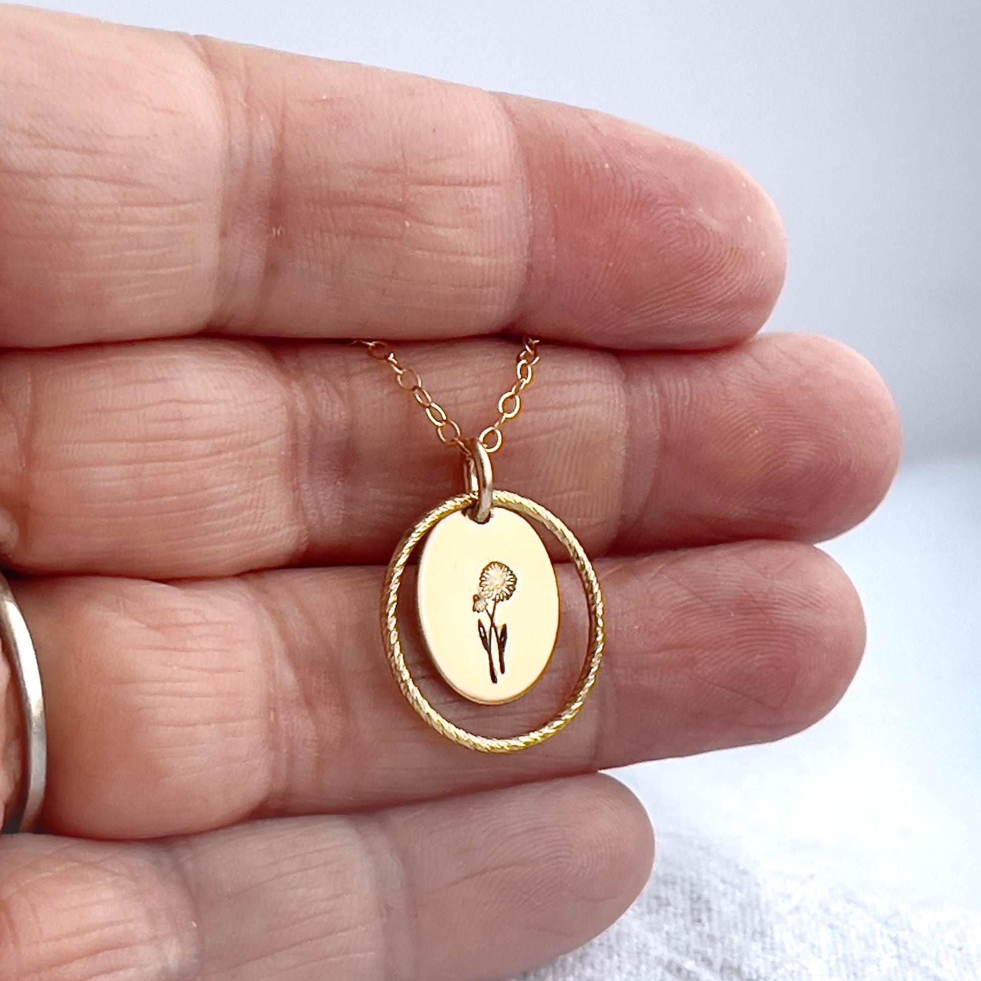 image of an oval gold birth flower necklace with a daisy stamped on it for april and a sparkle oval link surrounding it. the necklace is being held in a hand