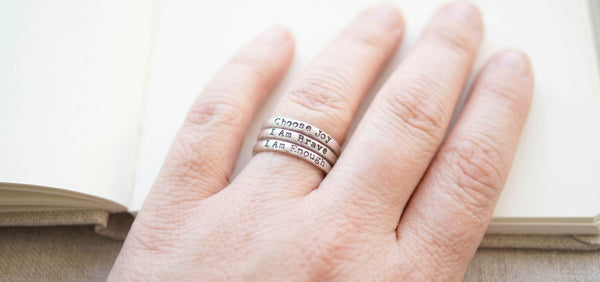 hand resting on a book wearing 3 stacking rings saying Choose Joy , I Am Brave, I Am Enough
