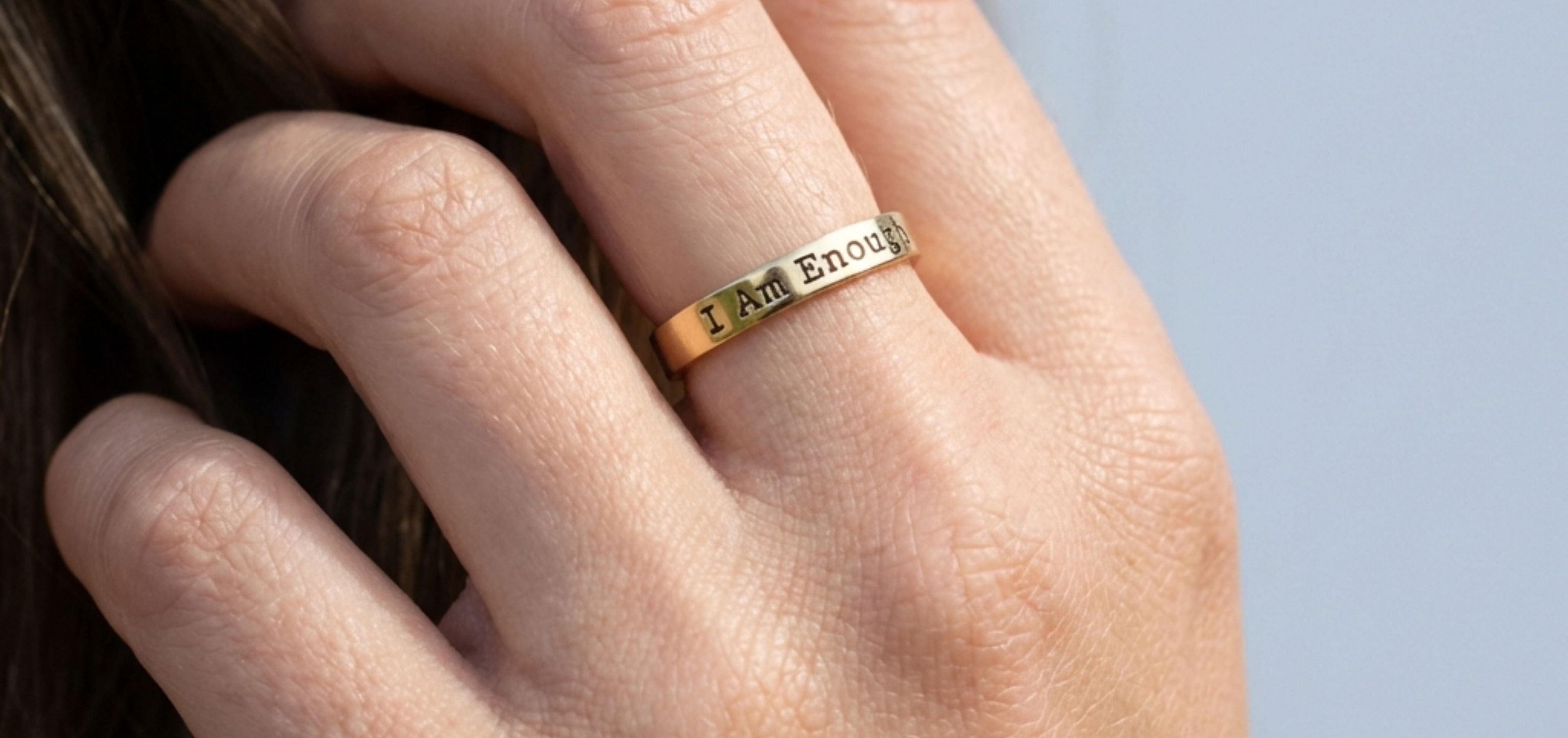 Close-up of a gold “I Am Enough” hand-stamped ring worn on a finger, promoting inspirational jewelry with meaningful quotes for confidence and self-worth.