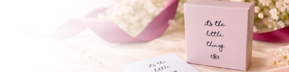 A light pink square gift box with black cursive text reading 'it's the little things' and a small lotus logo, placed on a white surface with soft floral props in the background.