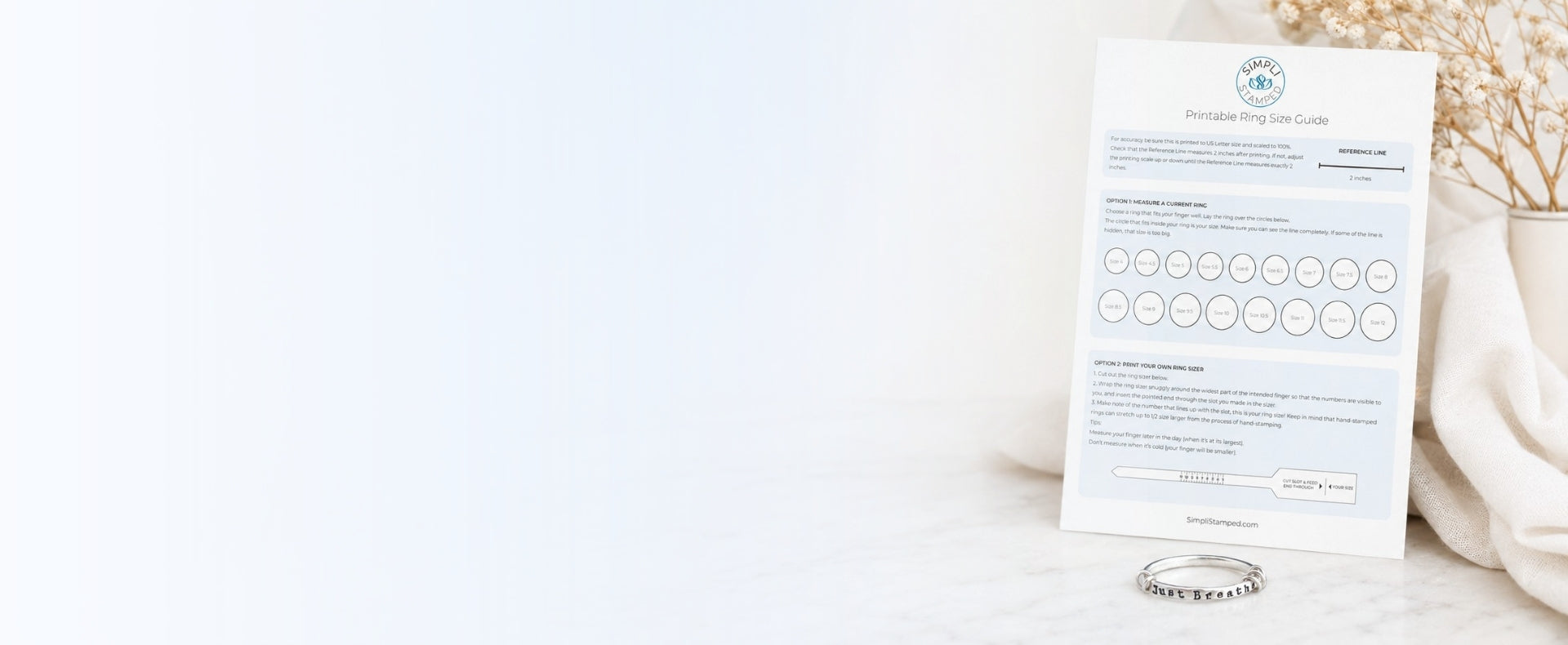 A printed ring sizing guide card with circular size options and a small silver just breathe ring placed beside it on a light marble surface, with soft neutral decor in the background.