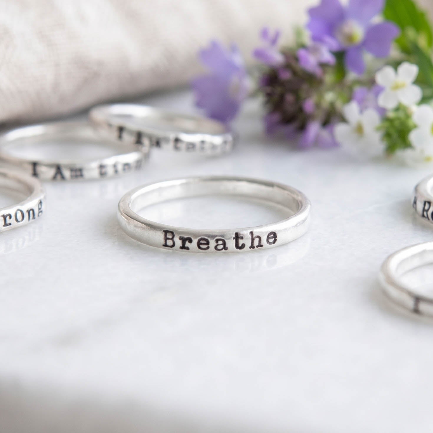 Custom Personalized choose your word ring in sterling silver stamped with Breathe on a white background with flowers and other rings behind.