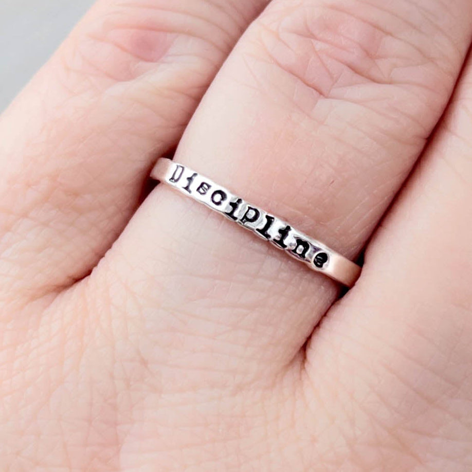 Hand wearing a silver ring that says Discipline on a neutral background