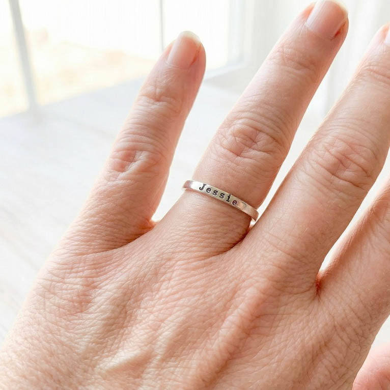 Hand wearing a silver ring with 'Jessie' stamped on it against a blurred background.