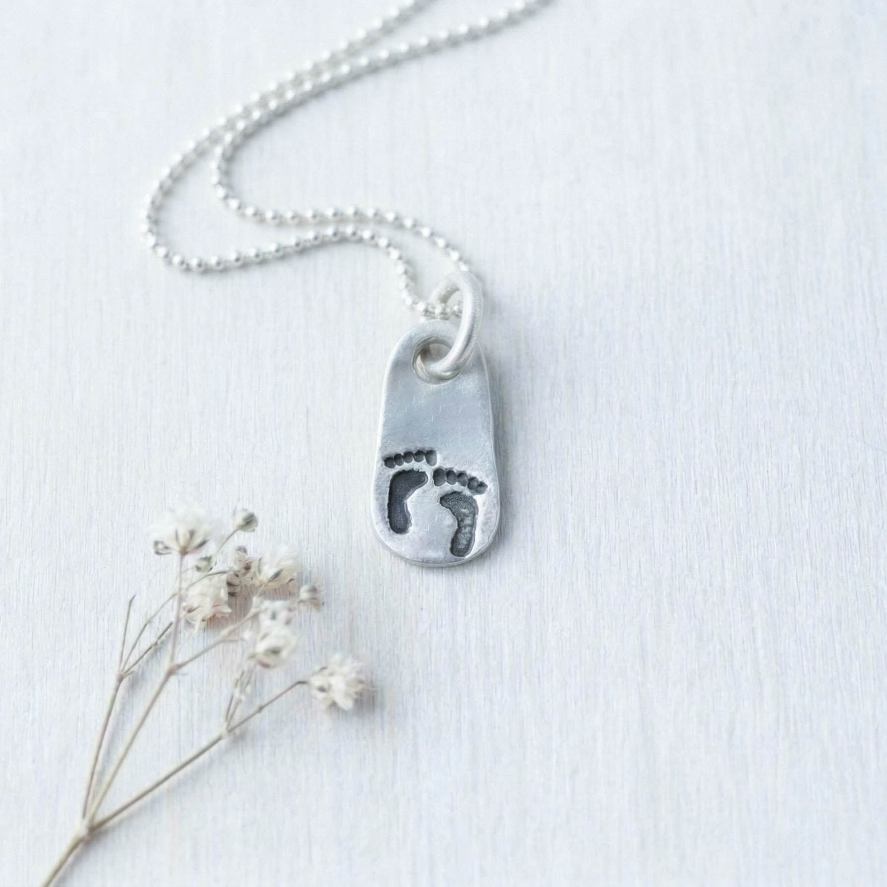 image of silver pendant with little baby footprints on a grey background with small flower below