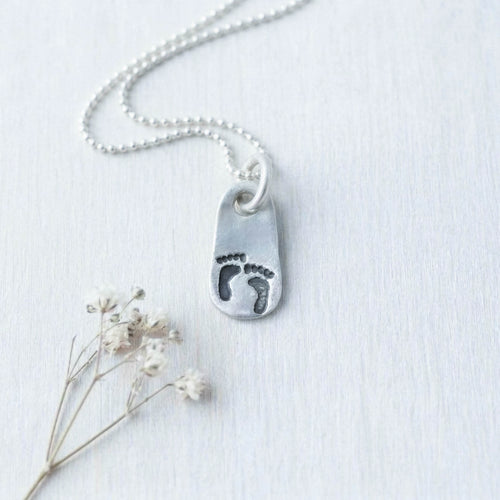 image of silver pendant with little baby footprints on a grey background with small flower below