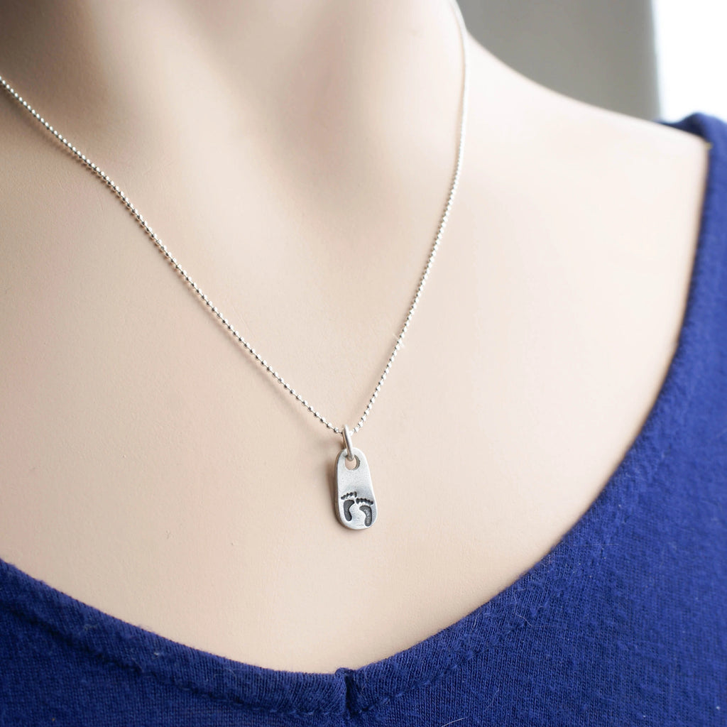 Silver necklace with a pendant with baby footprints on a mannequin wearing a blue top.