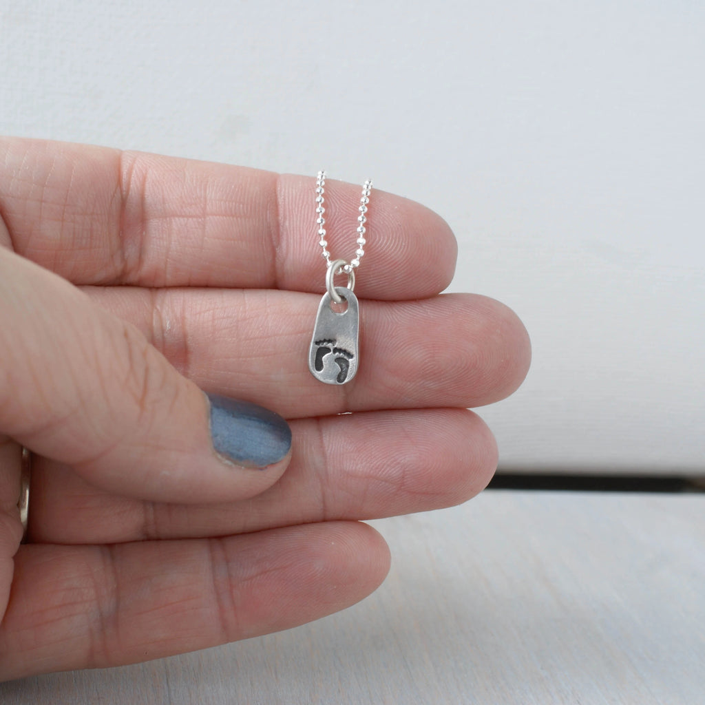 Hand holding a silver necklace with baby footprints on a small pendant against a plain background