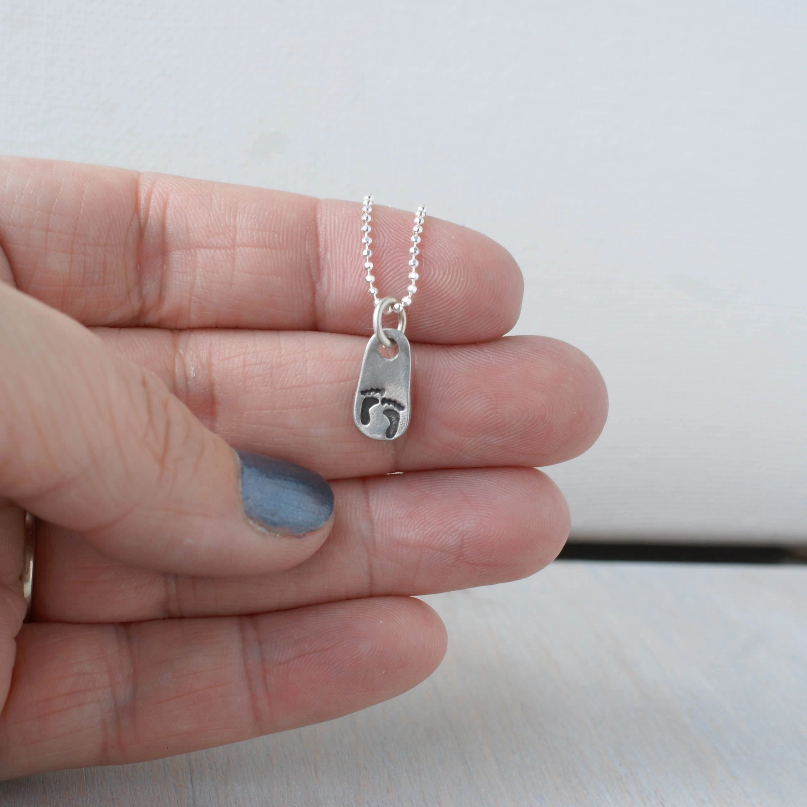 Hand holding a silver necklace with baby footprints on a small pendant against a plain background