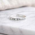 A sterling silver band ring stamped with the words 'I Am Enough' in a lowercase typewriter font, resting on a white marble surface.