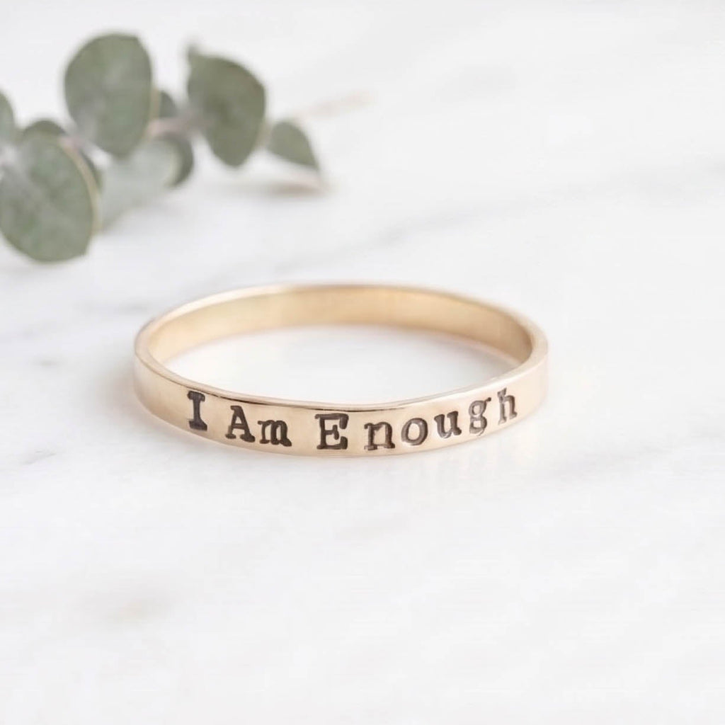 A slim gold band ring engraved with the words 'I Am Enough' resting on a white marble surface with blurred green eucalyptus leaves in the background.