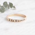 A slim gold band ring engraved with the words 'I Am Enough' resting on a white marble surface with blurred green eucalyptus leaves in the background.