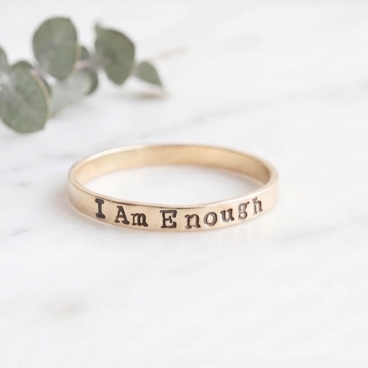 A slim gold band ring engraved with the words 'I Am Enough' resting on a white marble surface with blurred green eucalyptus leaves in the background.