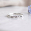 A silver band ring stamped with the words 'I Am Worthy' in sterling silver resting on a light gray reflective surface with a soft-focus background.