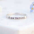 A silver band ring engraved with the words 'I Am Strong' in a dark metallic font, resting on a white marble block with small white and blue flowers in the background.