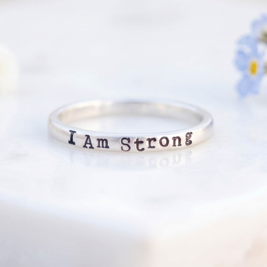 A silver band ring engraved with the words 'I Am Strong' in a dark metallic font, resting on a white marble block with small white and blue flowers in the background.