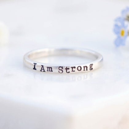 A silver band ring engraved with the words 'I Am Strong' in a dark metallic font, resting on a white marble block with small white and blue flowers in the background.