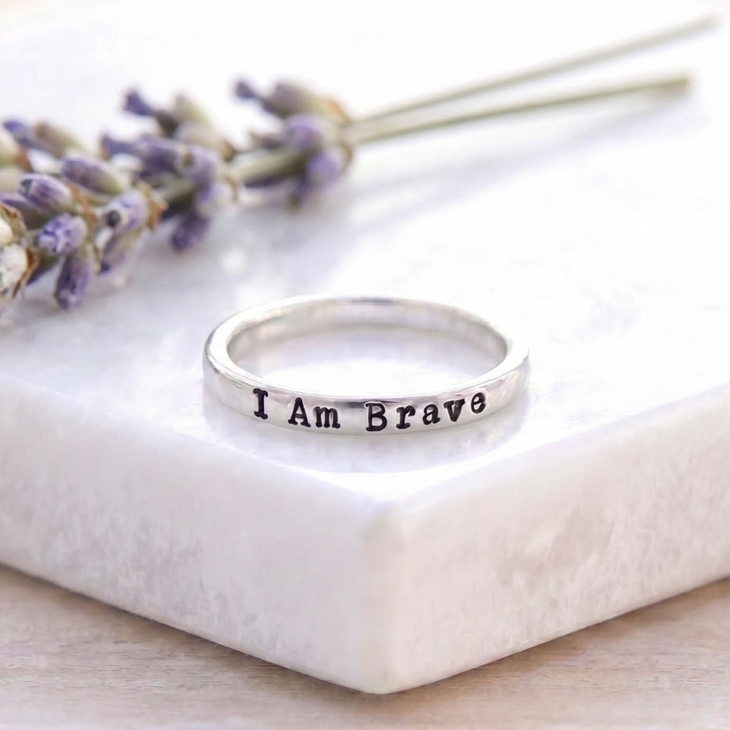 A silver band ring engraved with the words 'I Am Brave' in black lettering, resting on a white marble block with dried lavender sprigs in the background.