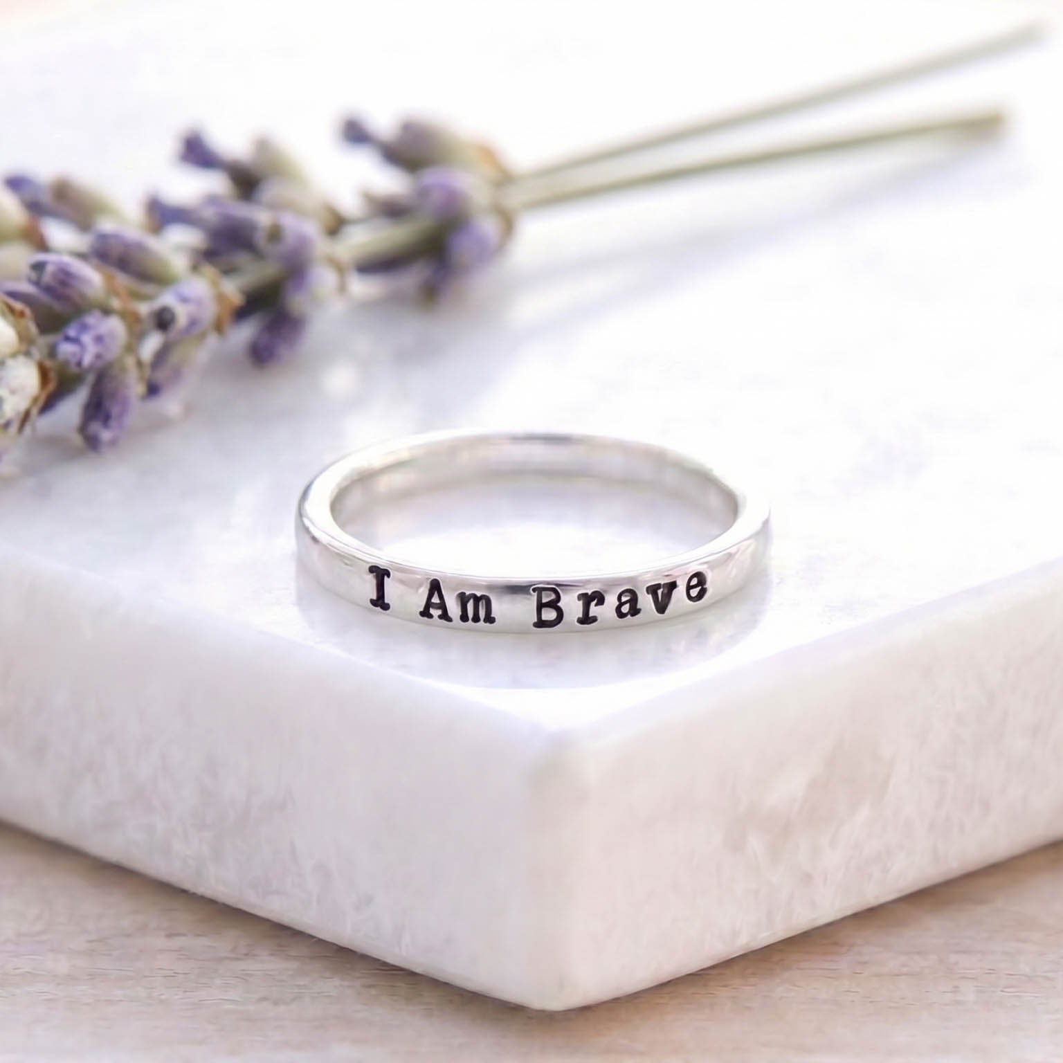 A silver band ring engraved with the words 'I Am Brave' in black lettering, resting on a white marble block with dried lavender sprigs in the background.
