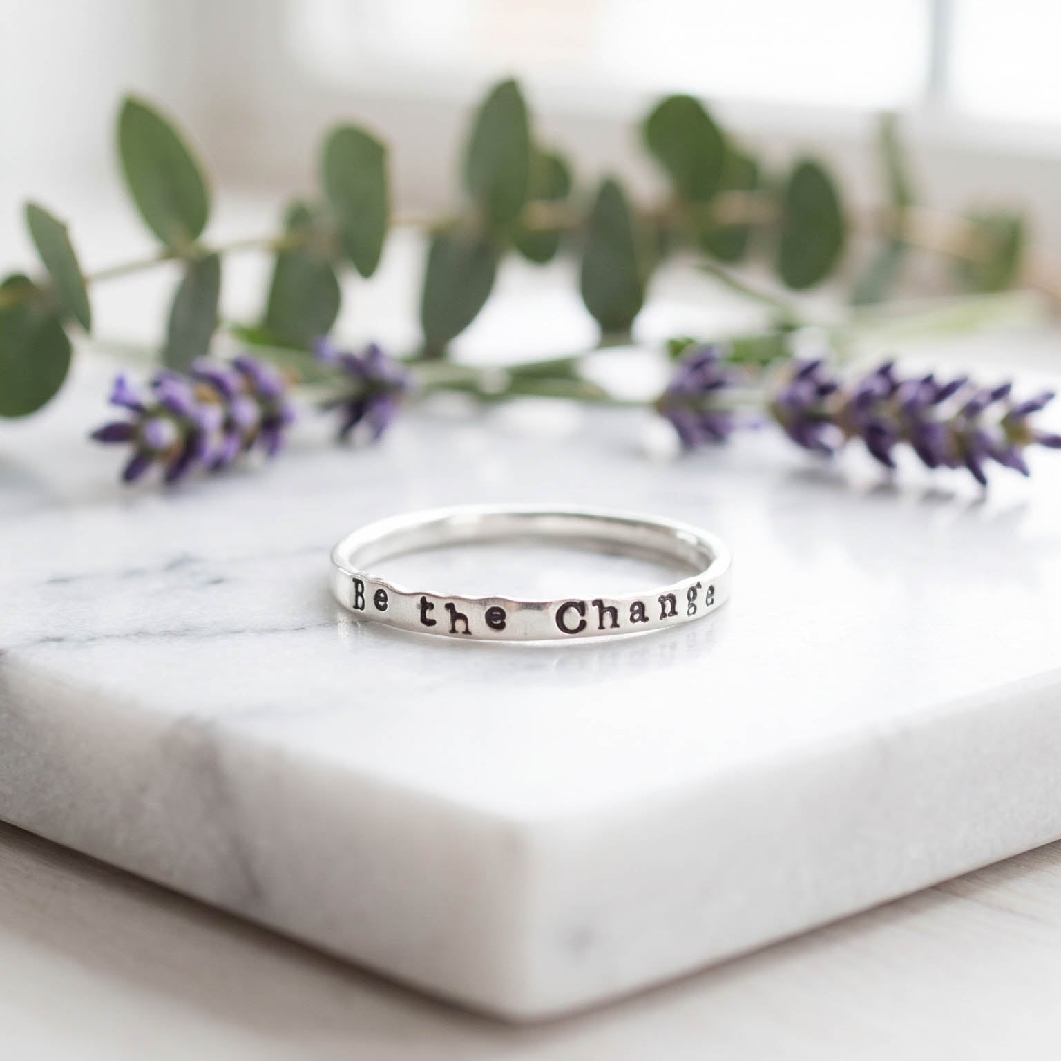 sterling silver ring stamped with Be the Change on marble surface with florals behind.