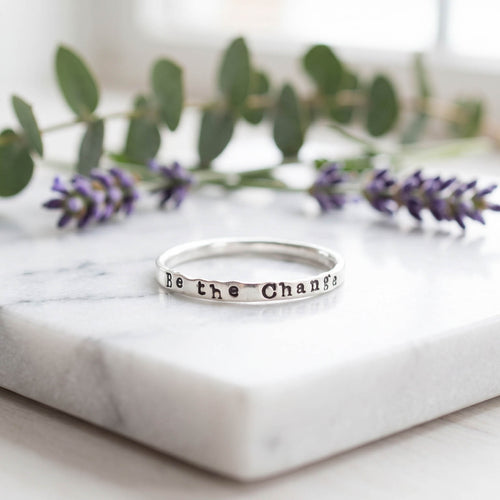 sterling silver ring stamped with Be the Change on marble surface with florals behind.