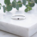 A thin silver band ring with a small black heart engraving, resting on a white marble block with blurred green eucalyptus leaves in the background.
