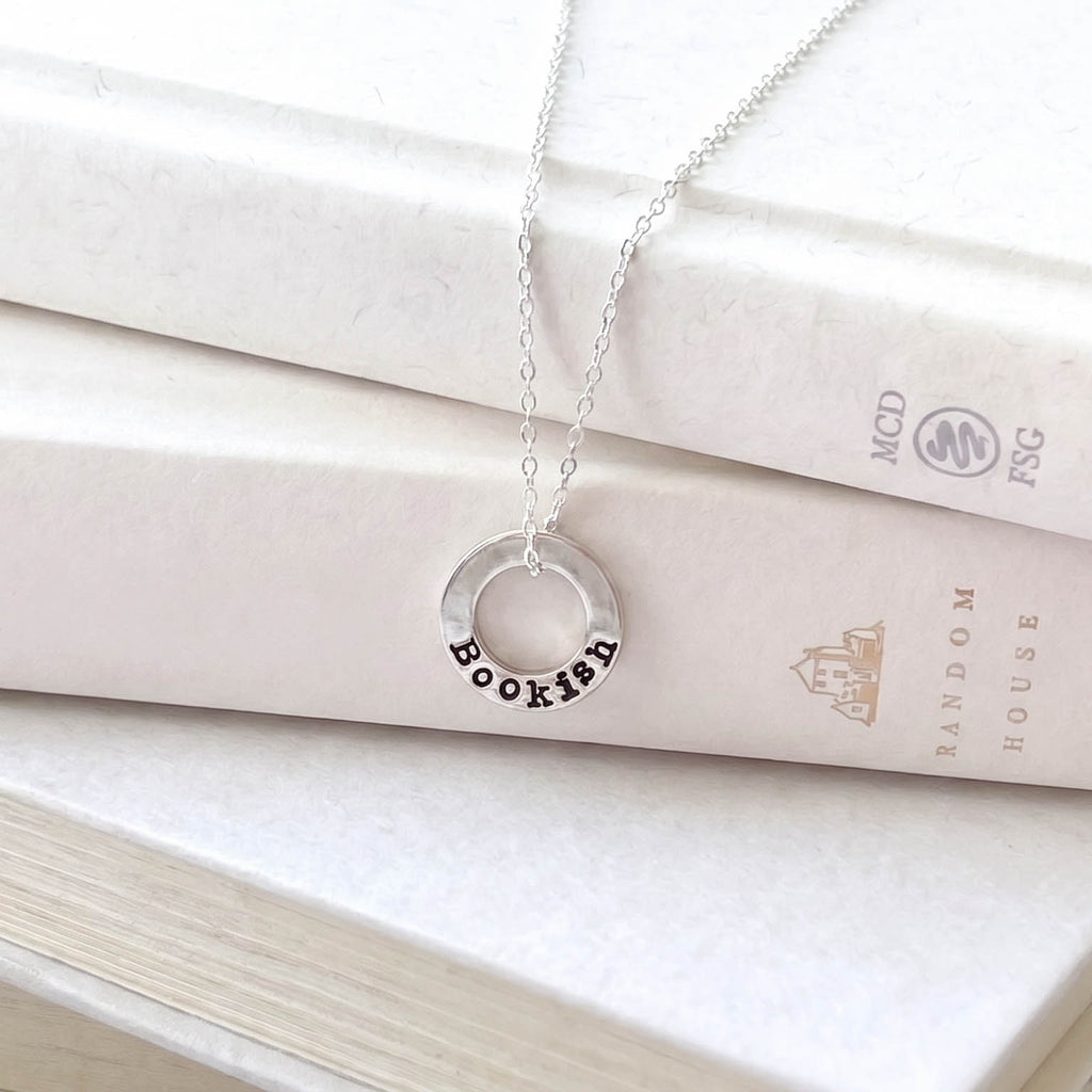 A silver necklace with a circular pendant engraved with the word 'Bookish' in black lettering, draped over stacked cream-colored books.