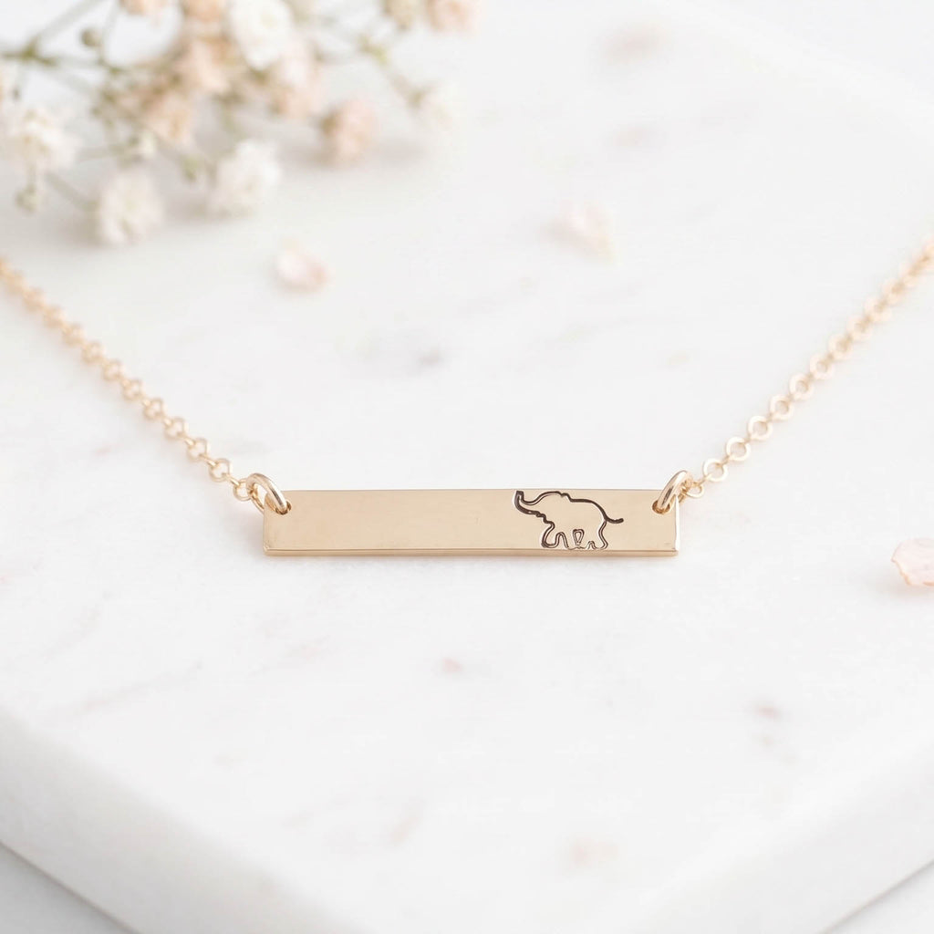 A delicate gold bar necklace with a small engraved elephant design, resting on a white marble surface with soft dried flowers in the background.