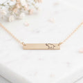 A delicate gold bar necklace with a small engraved elephant design, resting on a white marble surface with soft dried flowers in the background.