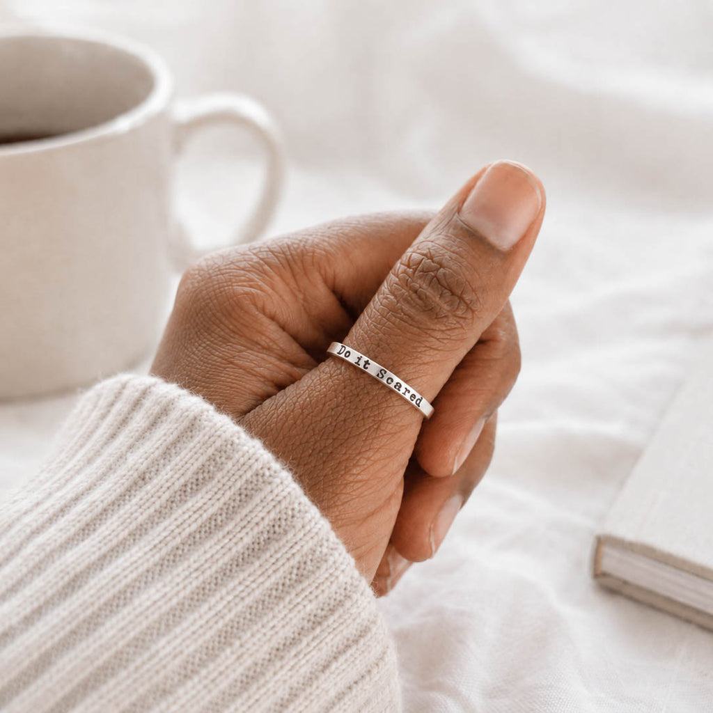 Hand wearing a ring with 'Do it Scared' on a blurred background with a cup and book.