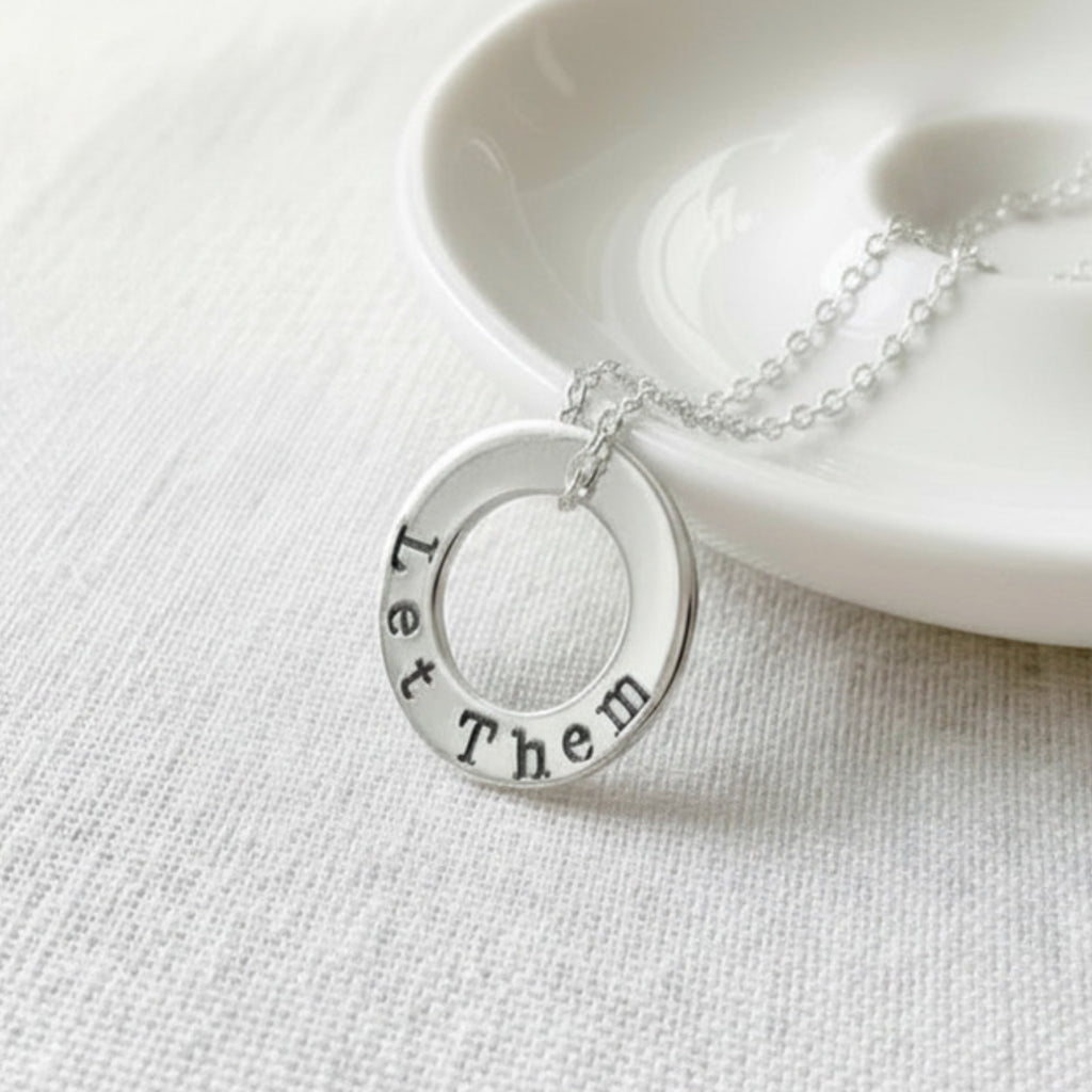 Silver washer necklace with Let Them stamped on it on a white surface