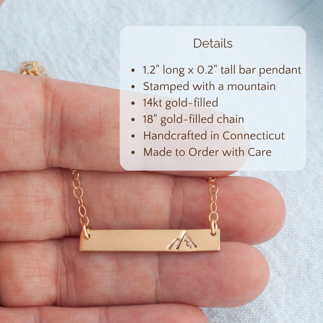 image of gold stamped bar with mountain held in a hand with product details overlayed. 1.2" long x0.2" tall bar pendant stamped with a mountain. 14kt gold filled.