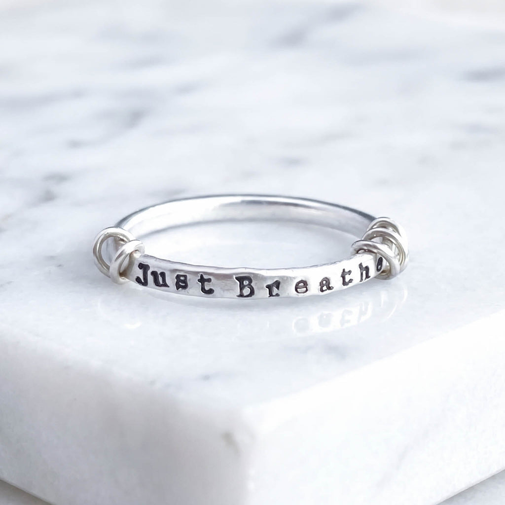 A sterling silver fidget ring with the words 'Just Breathe' hand-stamped on the front, resting on a white marble surface.
