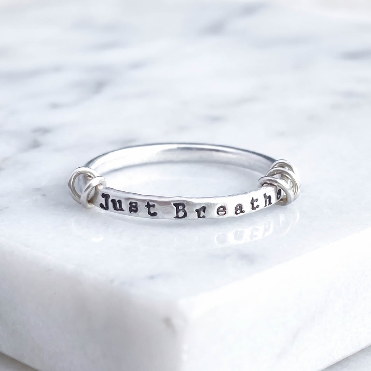 A sterling silver fidget ring with the words 'Just Breathe' hand-stamped on the front, resting on a white marble surface.