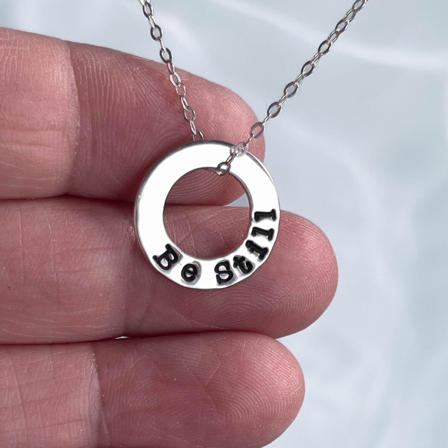 Silver necklace with circular pendant held between fingers against a white background