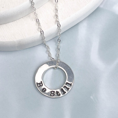 Silver necklace with a circular pendant engraved with 'Be Still' on a light gray background.