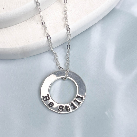 Silver necklace with a circular pendant engraved with 'Be Still' on a light gray background.