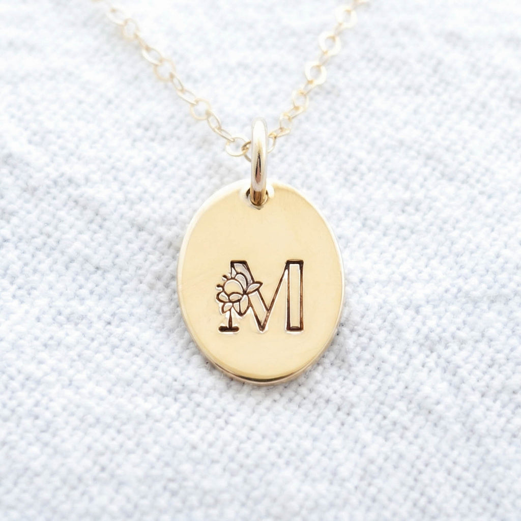 Gold filled floral initial necklace with an oval pendant on a light blue fabric background