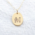 Gold filled floral initial necklace with an oval pendant on a light blue fabric background