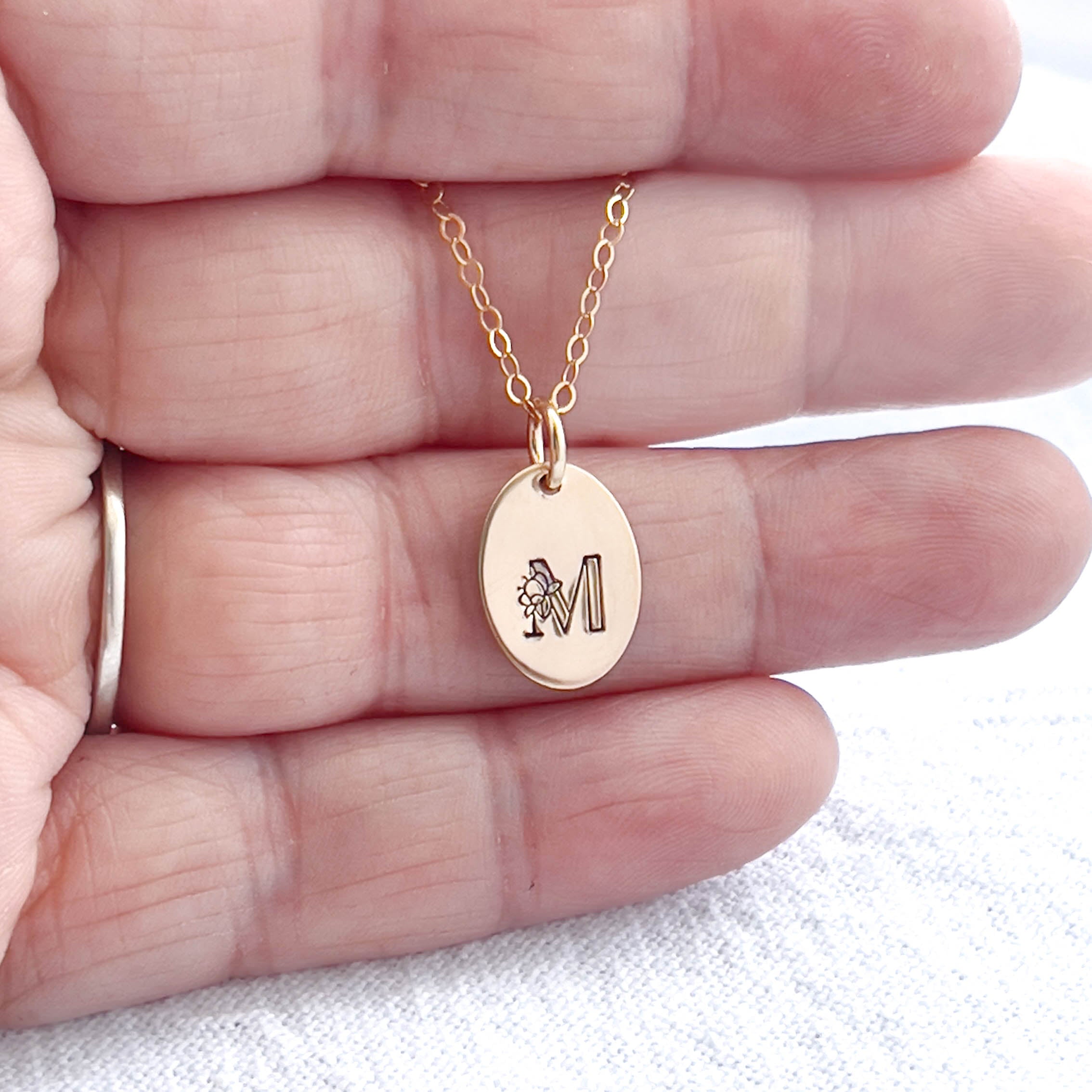 Gold necklace with a floral initial pendant featuring a letter 'M' held between fingers on a white background