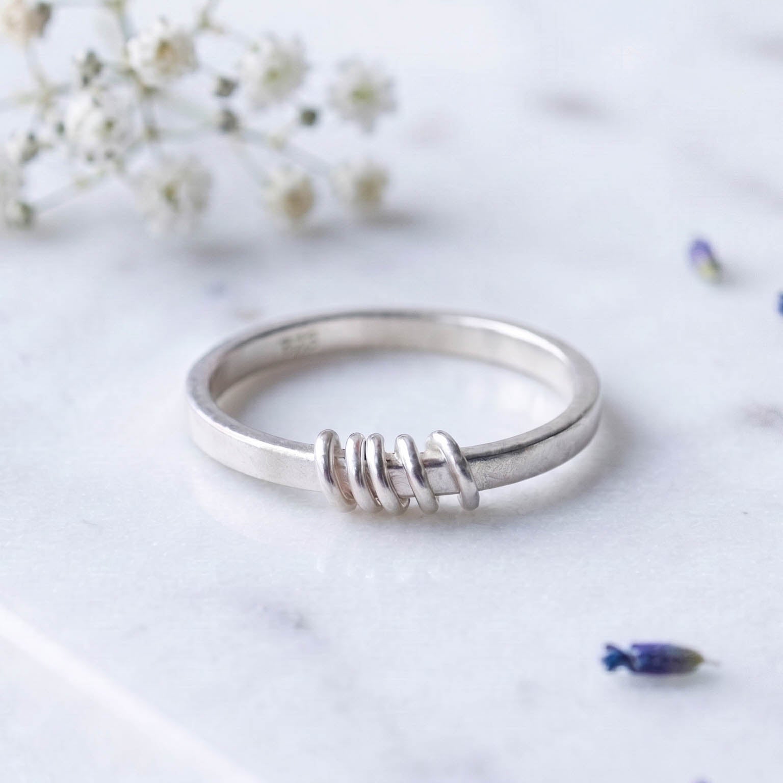 A silver spinner ring resting on a light marble surface with small white flowers and lavender buds in the background.