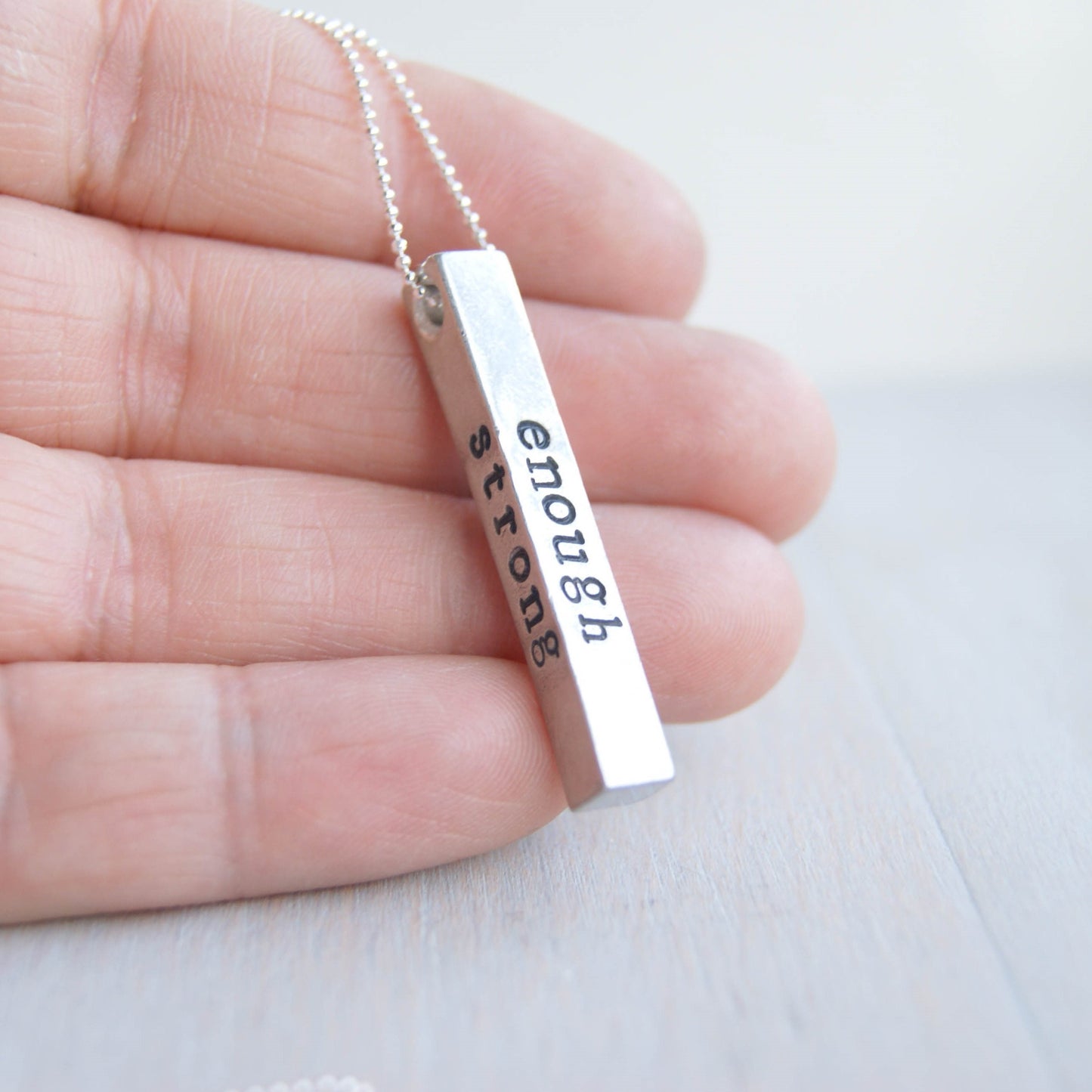 Long bar necklace stamped with strong, brave, and enough in artisan pewter held in hand