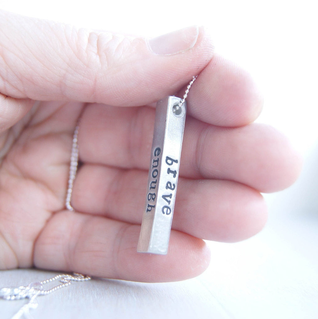 Long bar necklace stamped with strong, brave, and enough in artisan pewter held in hand