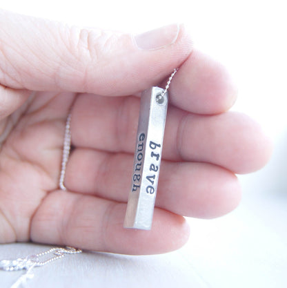 Long bar necklace stamped with strong, brave, and enough in artisan pewter held in hand