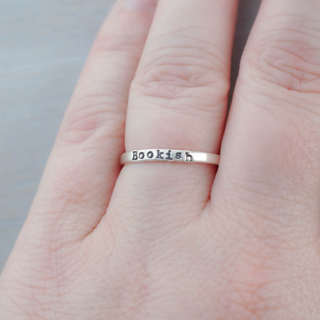 Silver ring with 'Bookish' engraving on a person's finger against a neutral background