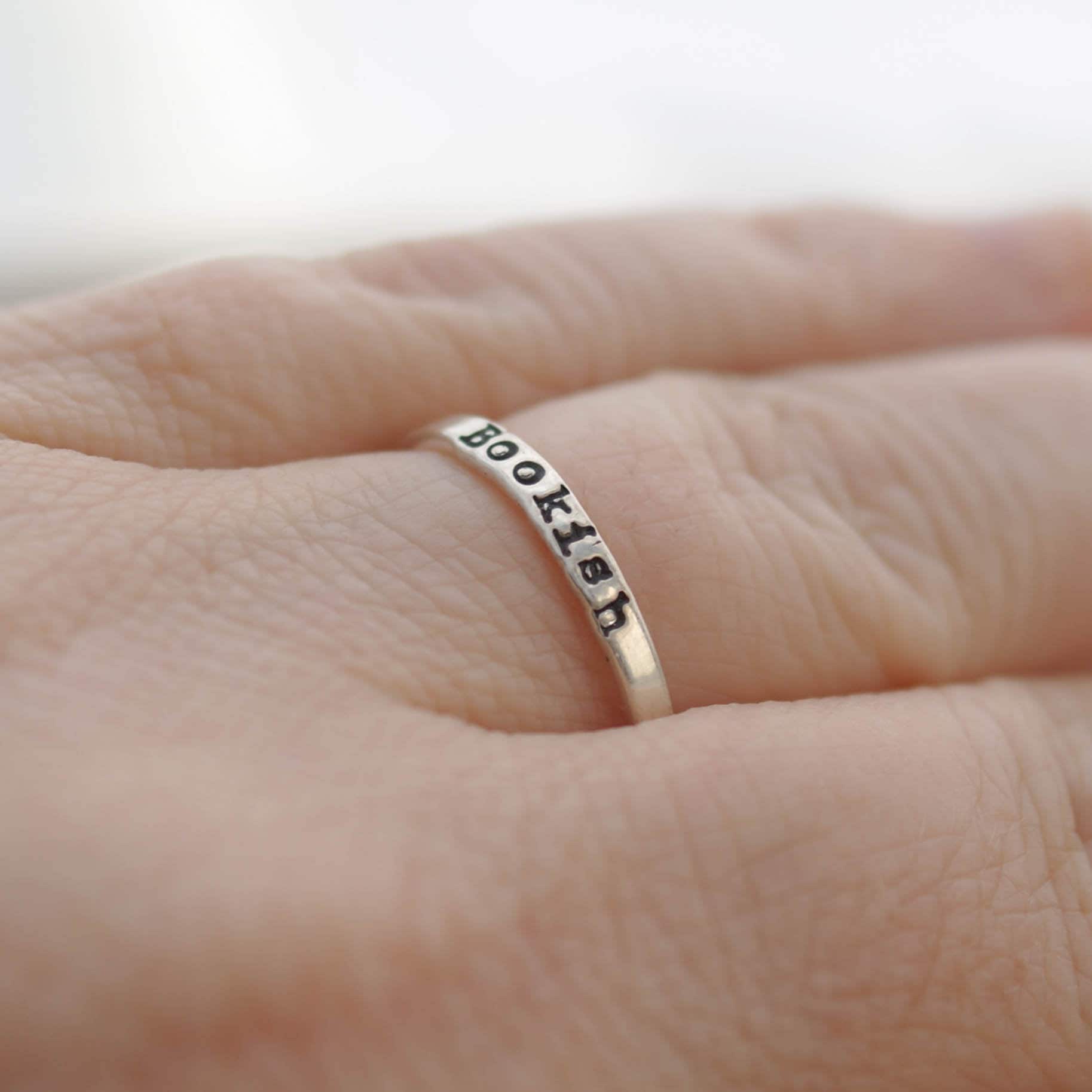Silver ring with 'BOOKISH' engraving on a person's finger against a neutral background