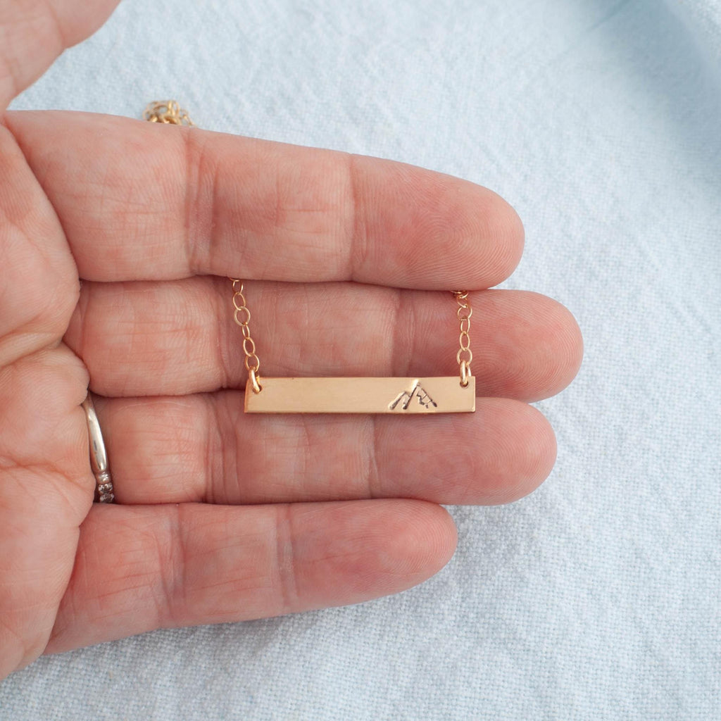 Image of gold necklace with mountain stamped on the right side being held in hand.