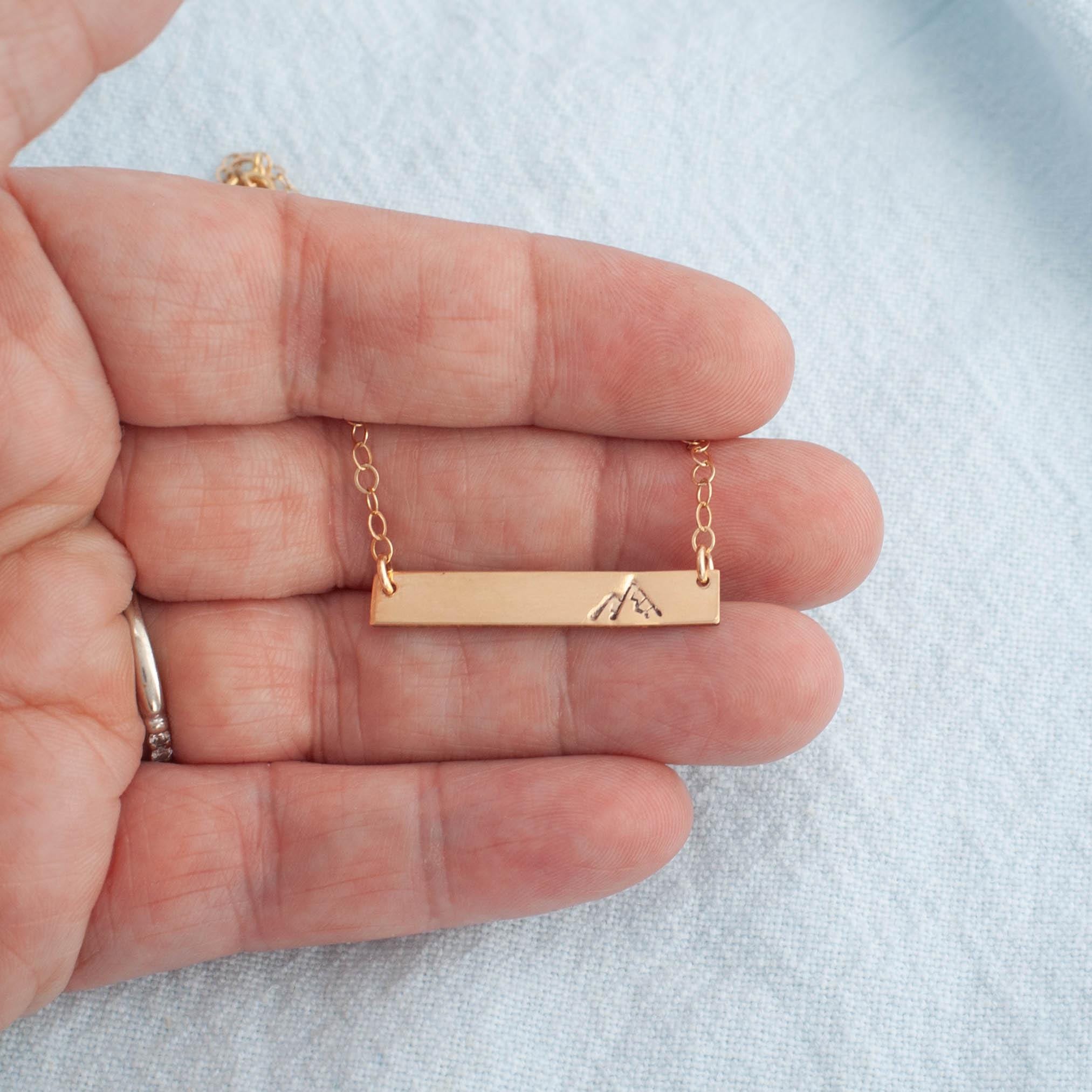 Image of gold necklace with mountain stamped on the right side being held in hand.