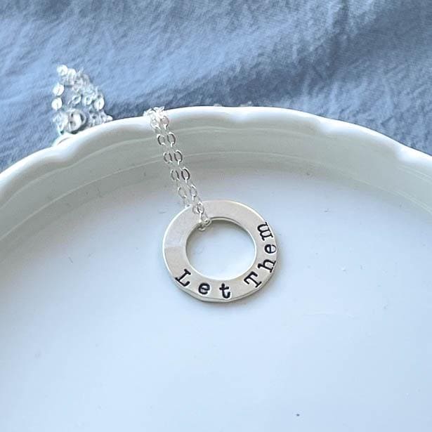 Sterling Silver Necklace stamped with Let Them on white tray with blue background