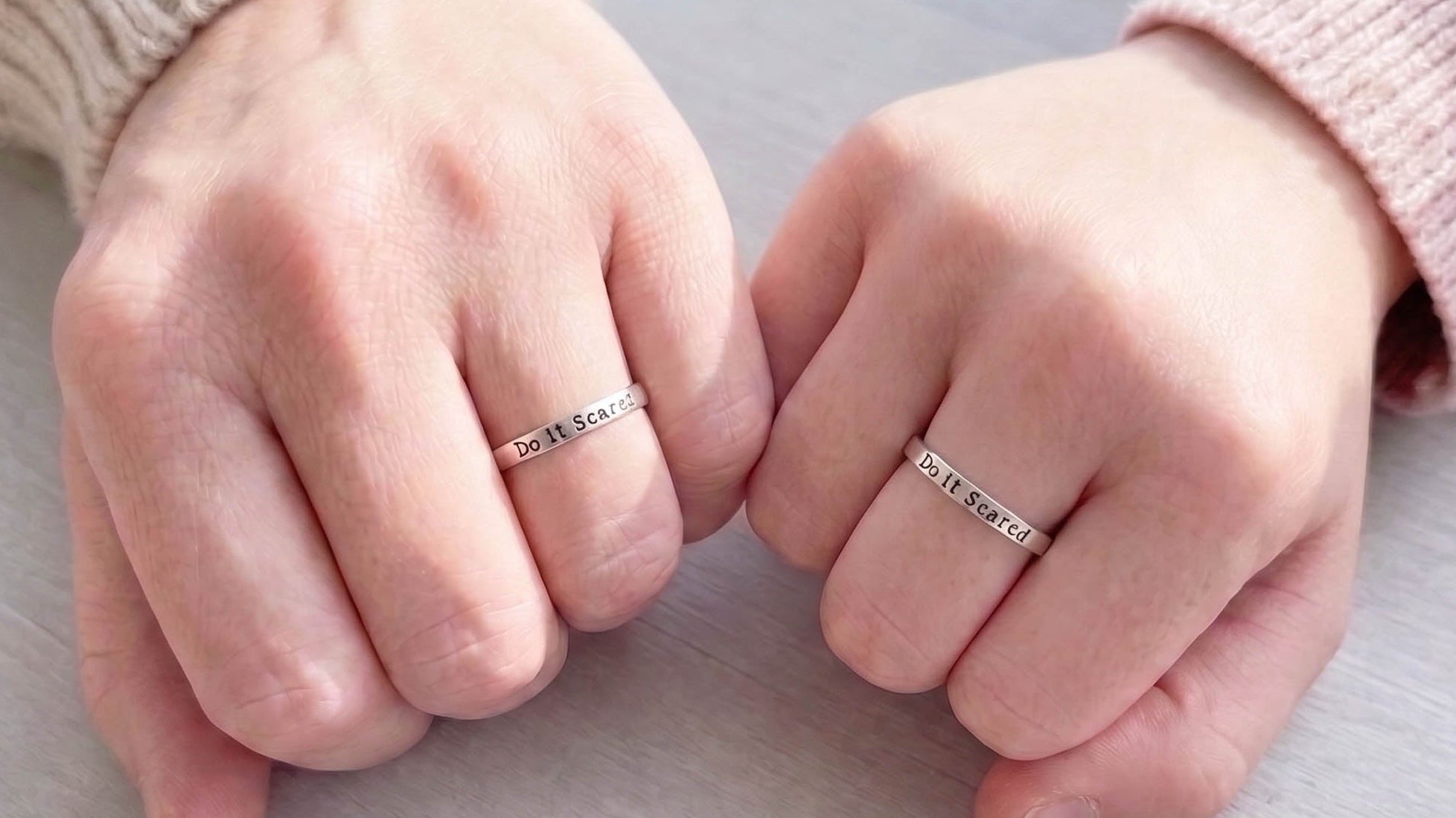 Two hands wearing slim silver rings stamped with Do it Scared. The person is wearing a cream ribbed sweater on one arm and a pale pink sweater on the other.